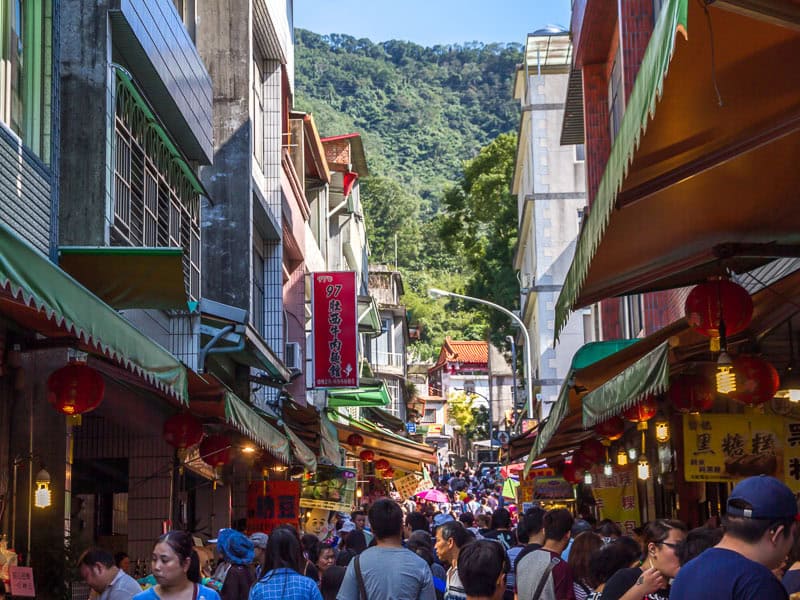 A crowd of people on a narrow street between buildings, with a small temple visible at the end