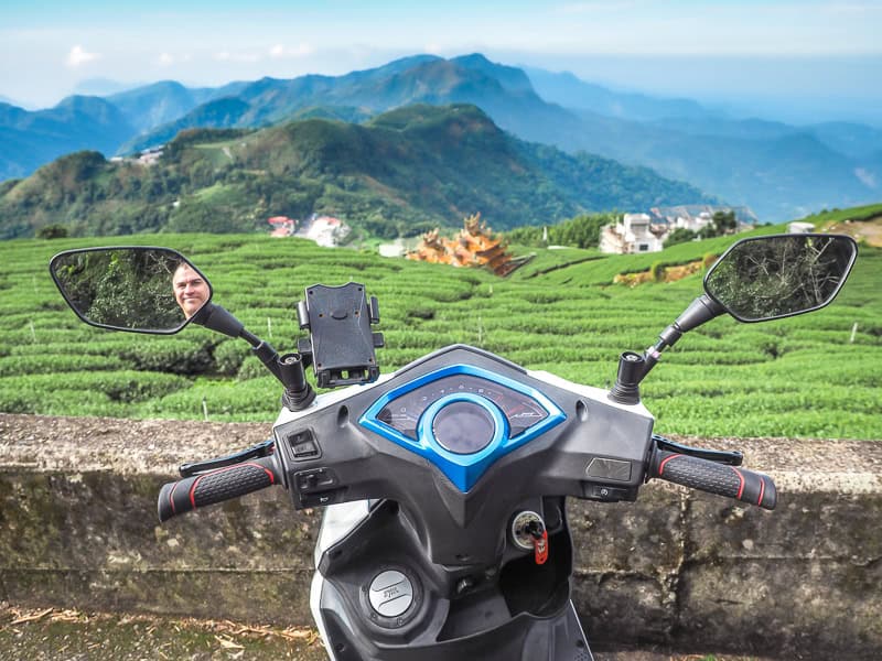 View looking down on some tea plantations, temple, and mountains, from the perspective of someone on a scooter, with Nick Kembel's face visible in the side view mirrow.