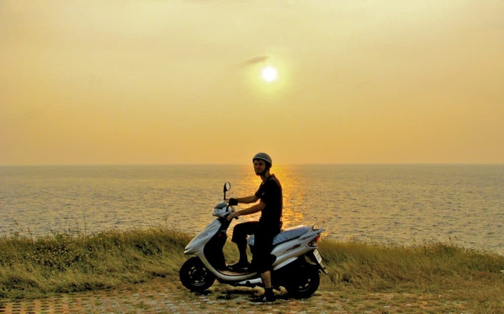 Nick Kembel sitting on a scooter on the coast, with sunset over the ocean behind him and the sky all yellow