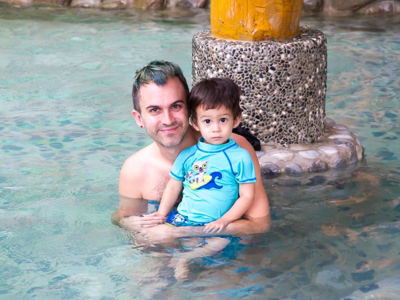 Nick Kembel with blue hair holding his son in blue bathing suit while sitting in hot spring water