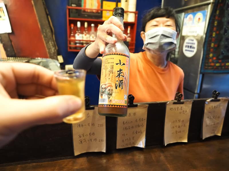 Nick Kembel holds out a shot of millet wine inside the photo, facing a Taiwanese aboriginal woman who is holding up the bottle it came from and smiling from behind a mask