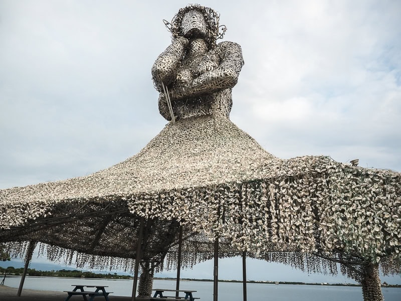 A large statue of a woman by the sea made of oyster shells