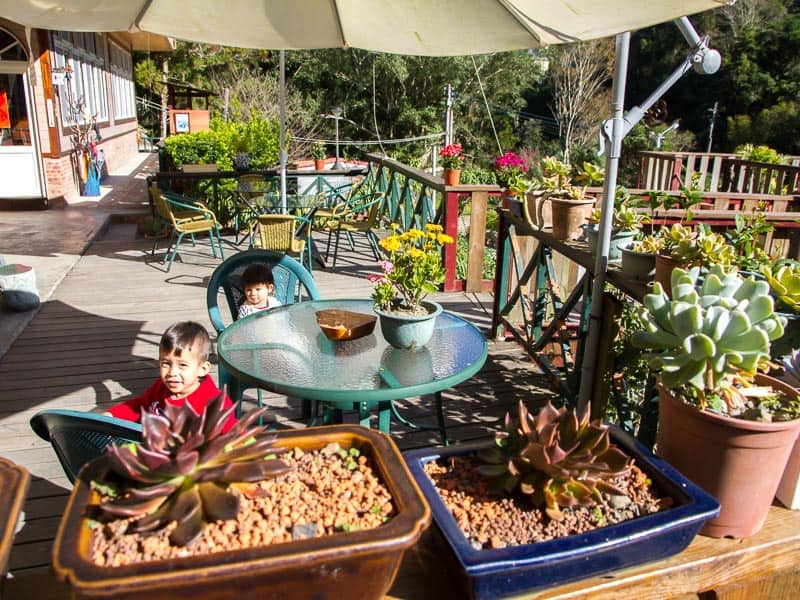 Two kids sitting at a round glass table on a large patio with plants