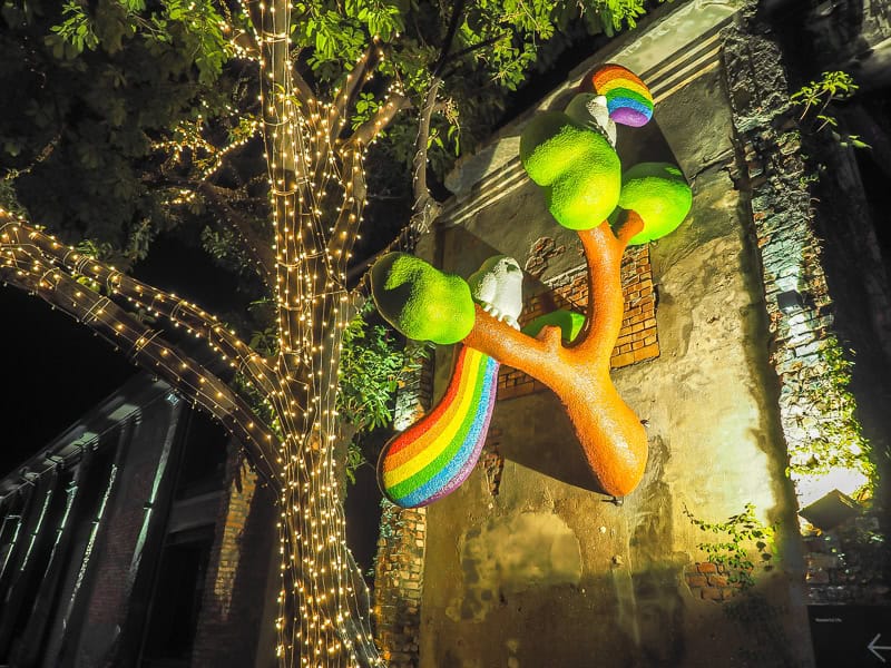 The side of a cement warehouse with some cute tree and bird statues sticking out from it and lights going up a tree