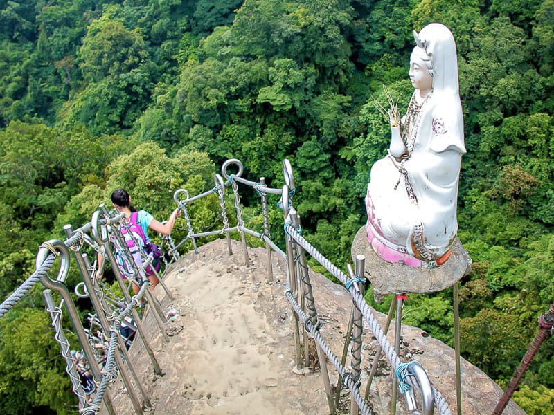 A hike descends from a vertical crag, shot from the top of the crag, with a Buddhist statue on the railing, and forest canopy down below
