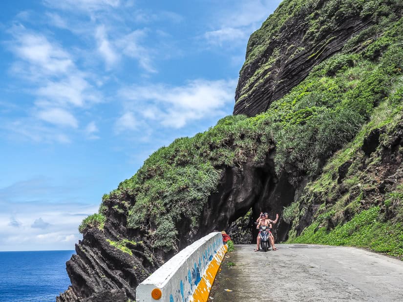 Two guys posing for the camera on a scooter on a small coastal road with cliffs down to the sea behind them
