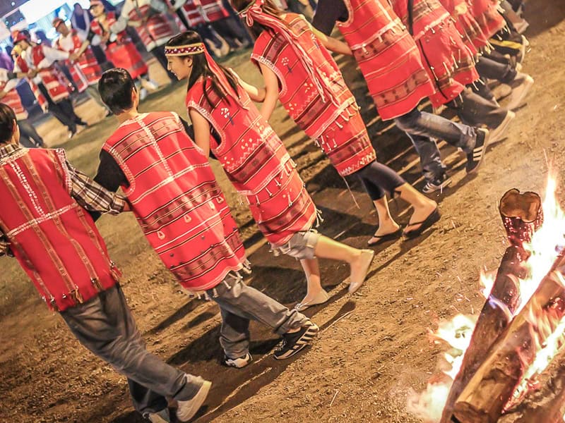 A large group of Taiwanese aboriginals wearing red costumes dance in a large circle beside a bonfire at night
