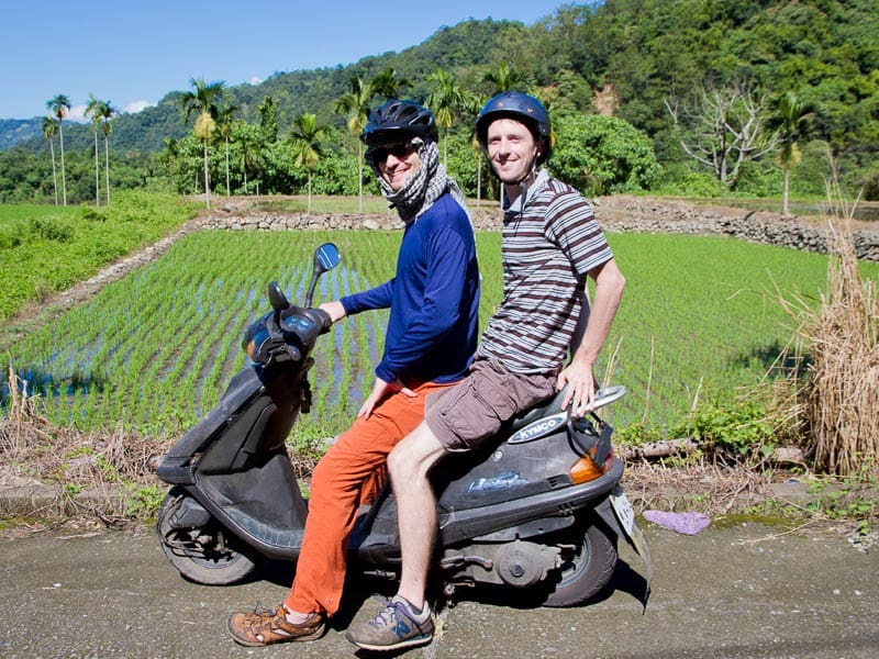 Two kids sitting on a scooter at the side of the road in the countryside, with rice fields behind them