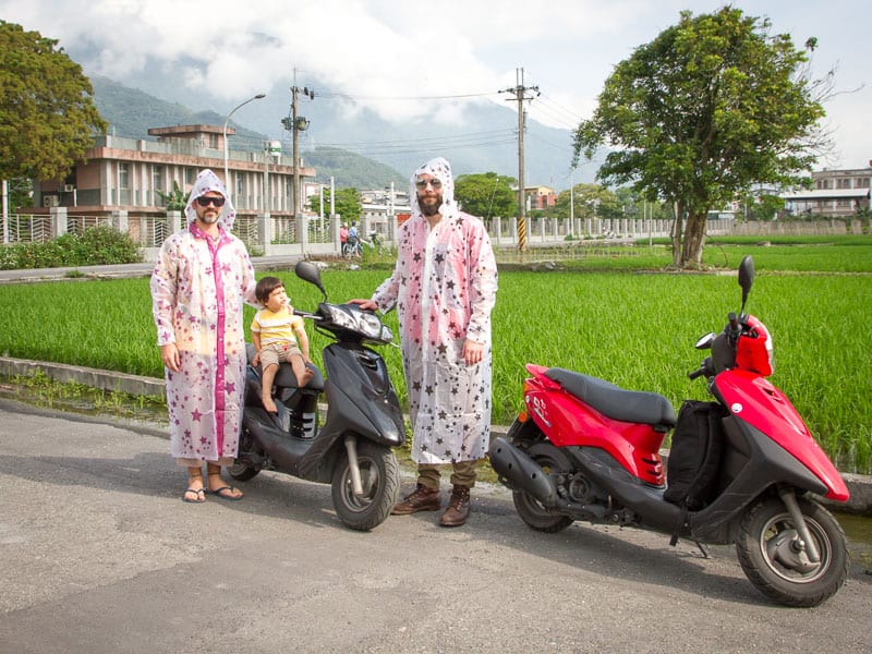 Two men standing in rain ponchos beside two scooters, next to a rice field, with a young buy sitting on one scooter and looking up at one of them