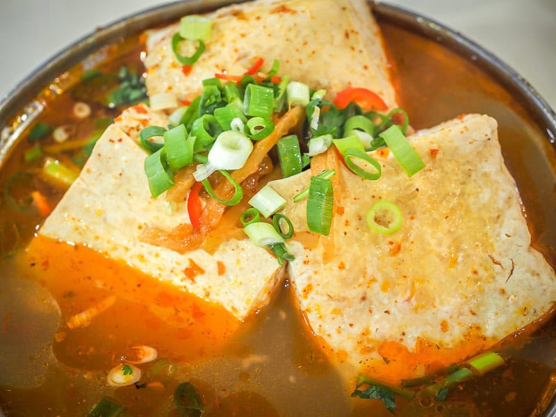 Close-up of a metal both of spicy broth with three big cubes of stinky tofu in it and garnished with diced green onions