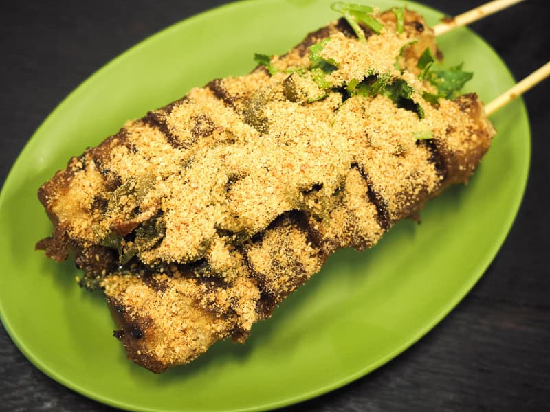 A green oval shaped plate with a log of grilled stinky tofu topped with copious peanut powder