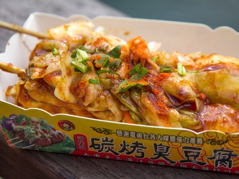 A paper tray with a log of grilled stinky tofu with many toppings and two sticks sticking out the end