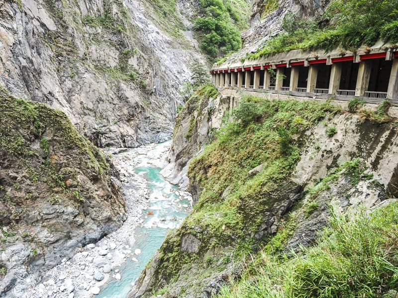 A small turquoise stream at the bottom of a valley and a car tunnel through the cliffs on the right