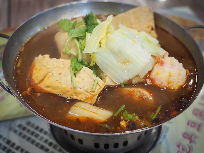 A metal pot with burner below containing several big chunks of tofu in a spicy soup with cabbage and cilantro on top
