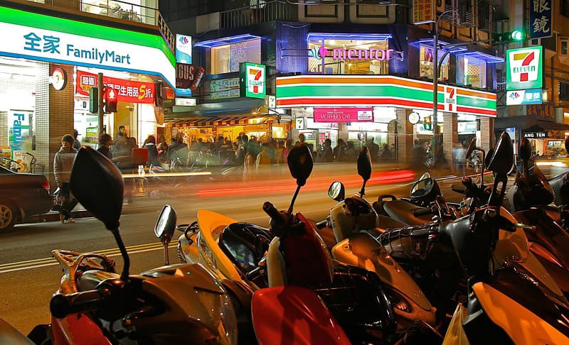 Two convenience stores side by side at night, with the blurs of passing traffic
