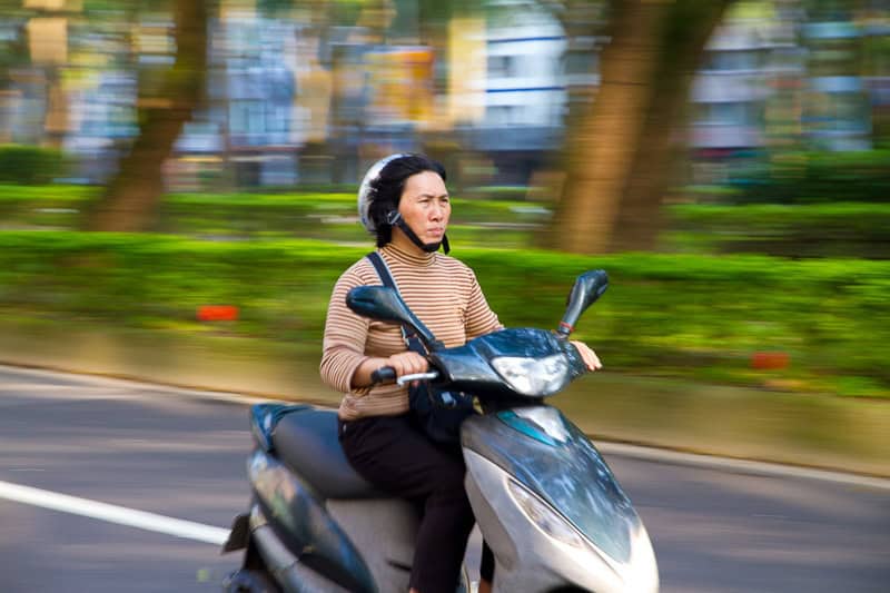 A Taiwanese middle aged woman riding a scooter in Taipei, with the background blurred due to the movement