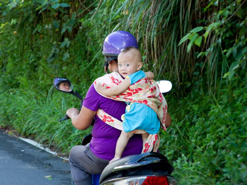 A Taiwanese woman riding a scooter, shot from behind, with a baby attached to her back with a scarf, and the baby is turning around to look at the camera