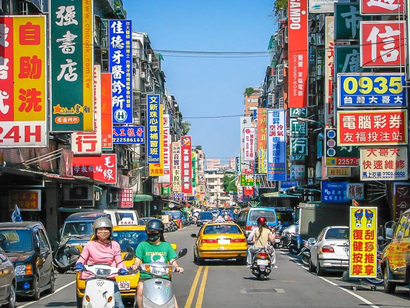 A typical street scene in Taiwan, with some people on scooters waiting at an intersection and many shop signs on either side of the road