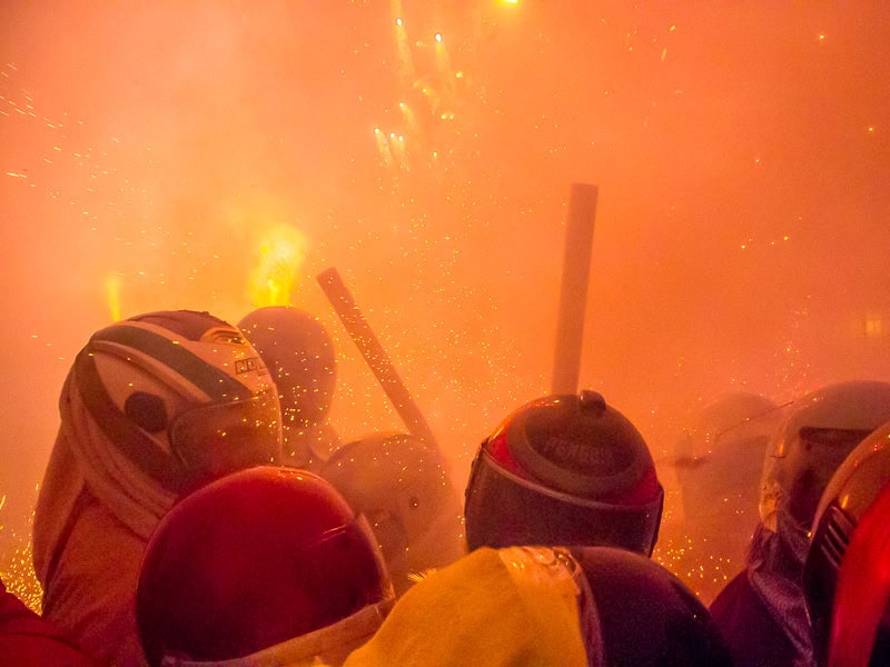 A dense crowd of people wearing helmets with the orange blaze of fireworks going off around them