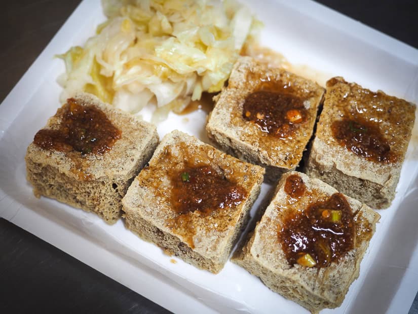A white tray with five cubes of deep fried tofu, each with a hole in it filled with spicy sauce, and some pickled vegetables on the side.