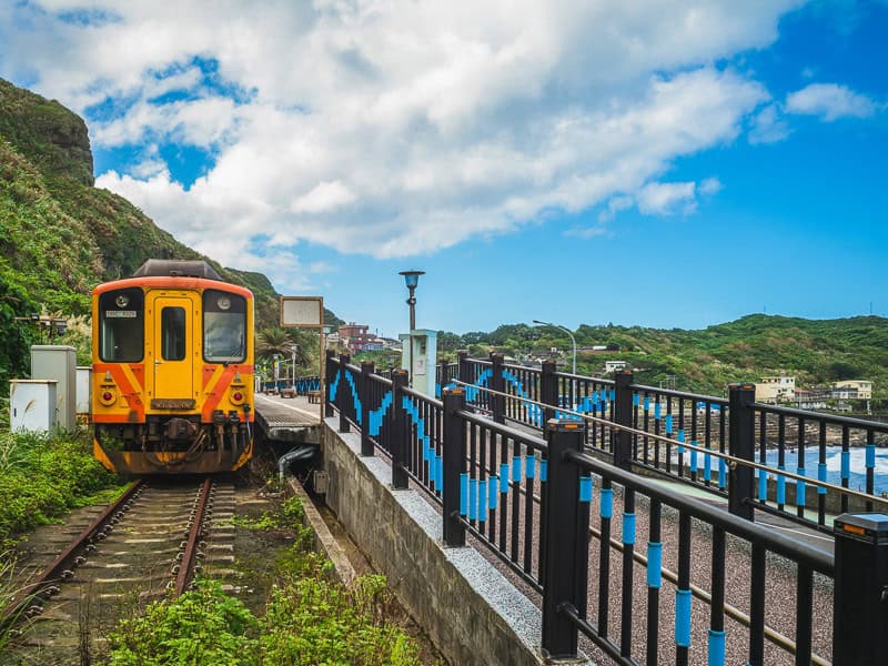 A yellow train or the Shen'ao Line parked at a platform with hills at the sea visible to the side