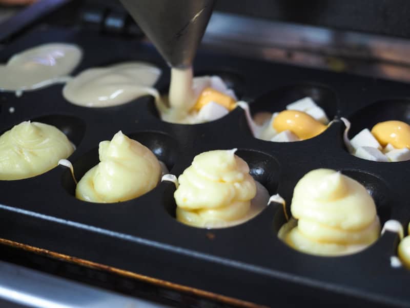 Close up of a metal rack with 10 slots filled with dough and a piping bag putting some cheese sauce into them