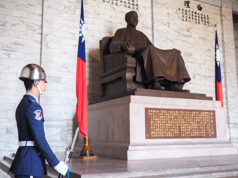 A uniformed guard stands beside a bronze seated statue of CKS with Taiwan flag beside