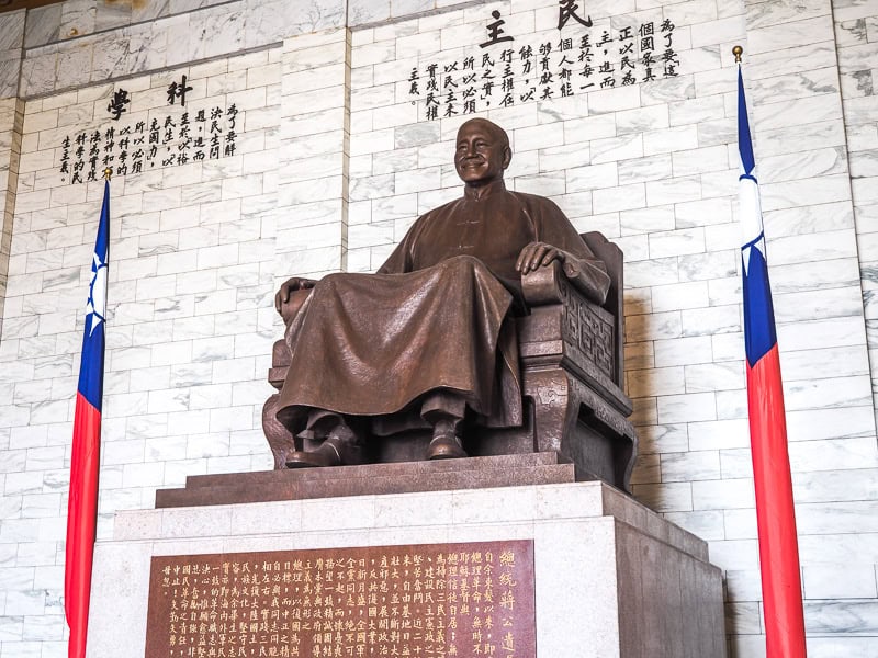 An elevated, bronze statue of a seated Chiang Kai Shek with Taiwan flag on the side and Mandarin characters on the white stone wall behind