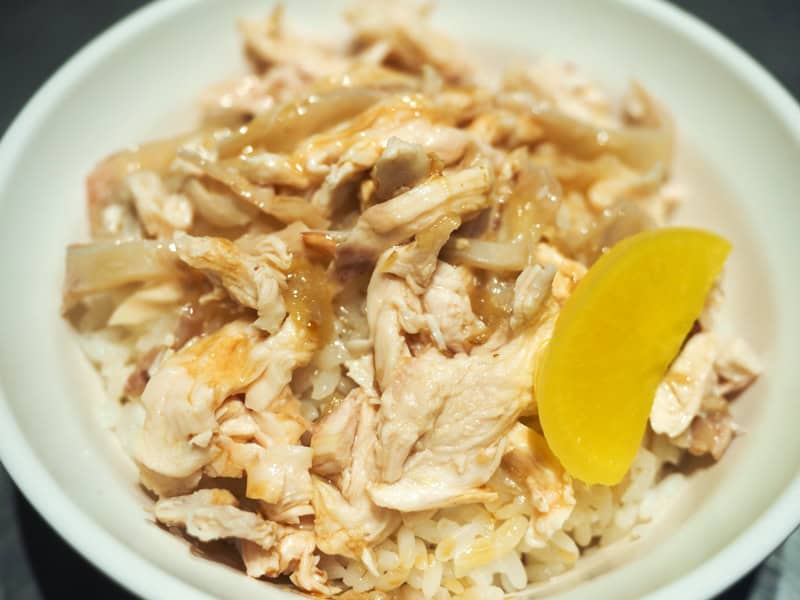 Close up of a bowl of rice topped with turkey meat, meat drippings, and a slice of bright yellow pickled radish