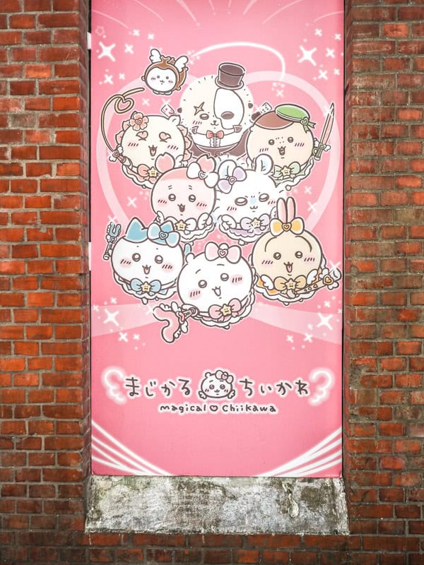 A tall skinny pink banner with Chiikawa anime characters on it hanging on a red brick wall