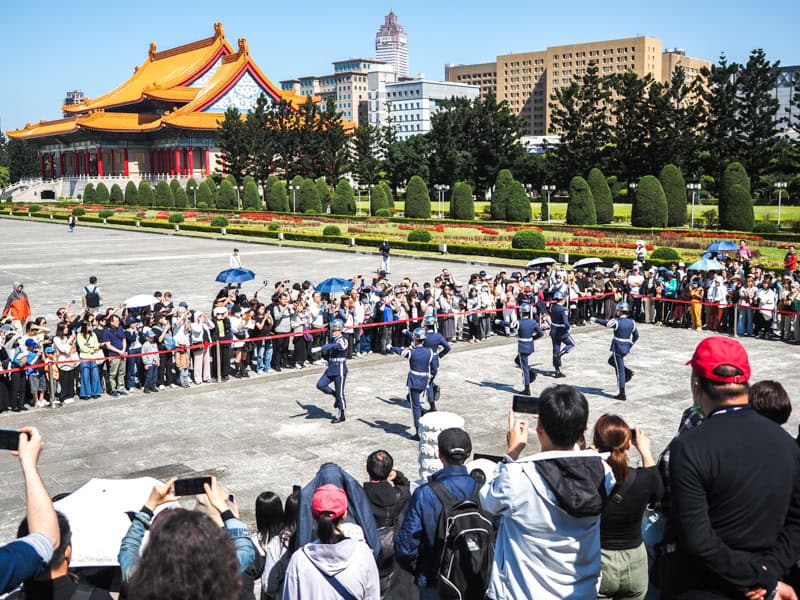 A crowd of people around a roped off section of a square with guards doing a changing ceremony