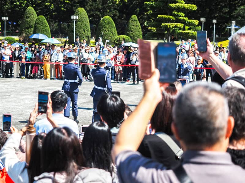 A crowd of tourists shot from behind as several hold up their phones to take photos of the CKS guard changing ceremony