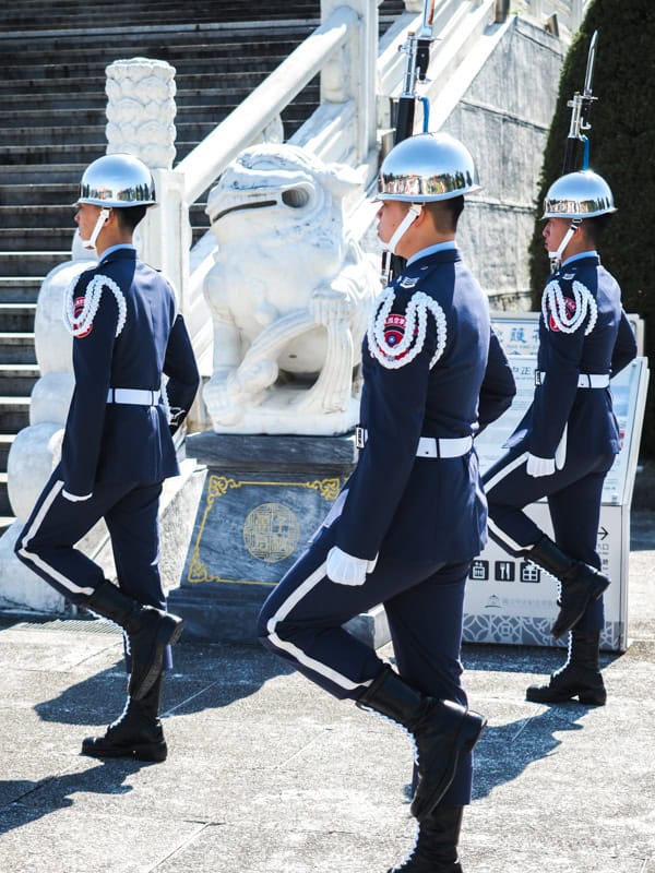 Three uniformed guards march past a stone lion to Liberty Square in front of CKS Memorial Hall