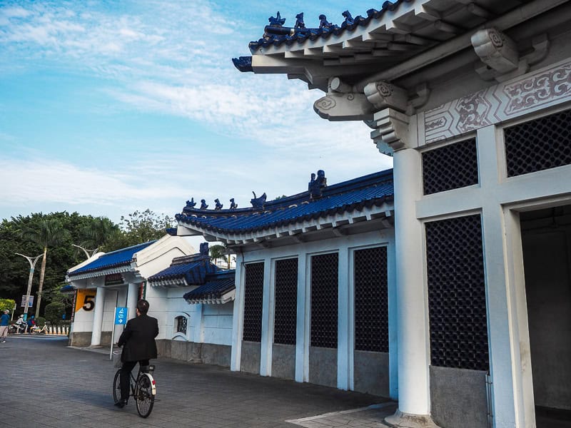 A man riding a bicycle past a traditional white gate with blue roof tiles and exit of CKS Memorial Hall MRT station