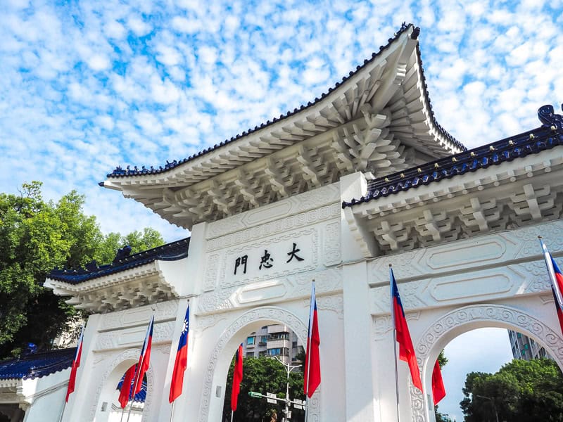 Looking up at one of the smaller white and blue gates providing access to Chiang Kai Shek Memorial hall, with several Taiwan flags standing at the bottom