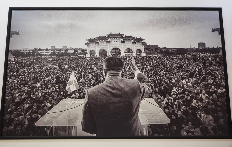 A large black and white photo of a speaker shot from behind as he delivers a speech to a huge crowd of people in Liberty Square at Chiang Kai Shek Memorial Hall in Taipei