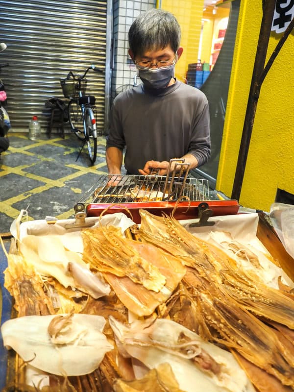 Vertical image of a middle aged Taiwanese man wearing mask behind a food stall, with the stall covered in a pile of different size full dried squids, and the man grilling one of them on a rack over a small fire
