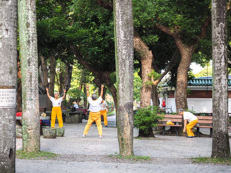 A group of elderly women in yellow pants and white shirts doing exercises between some trees on the CKS Memorial Hall grounds