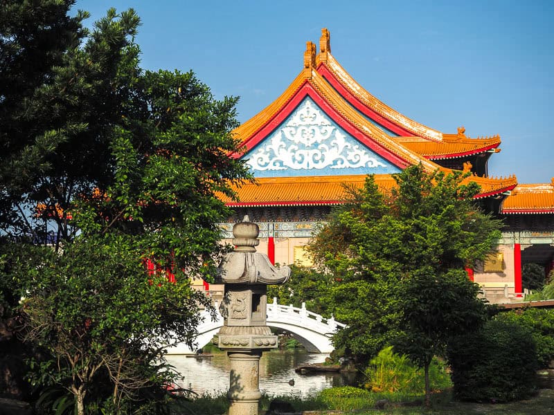 A pond with stone lantern, white arched bridge, and classical Chinese National Concert Hall behind