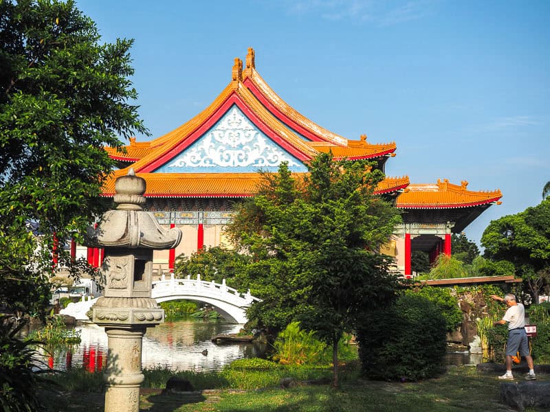 A man doing taichi at the side of a pond with stone lantern, white arched bridge, and National Concert Hall behind