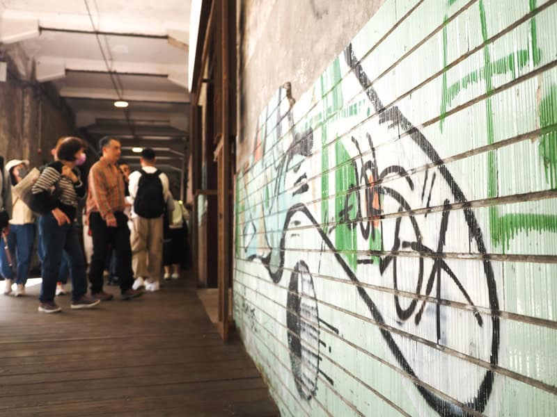 Close up of some spray painted art on a white brick wall, with some crowds of people entering a shop behind