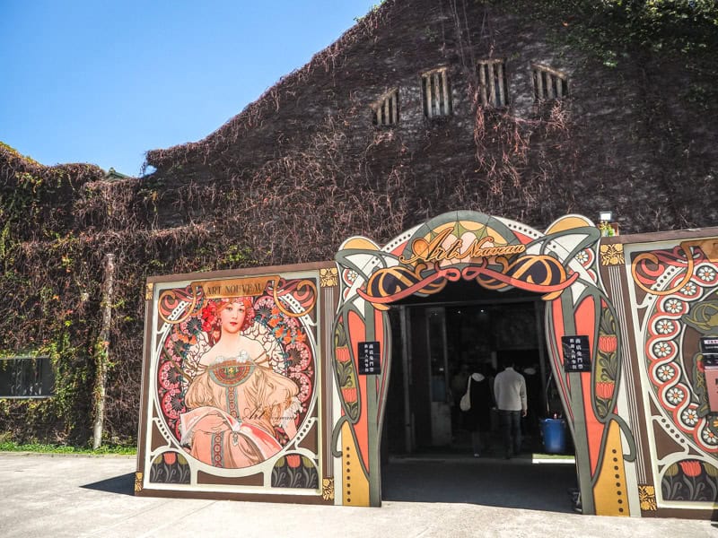 An old building covered in vines, with an entrance and large sign with an art nouveau painting on it