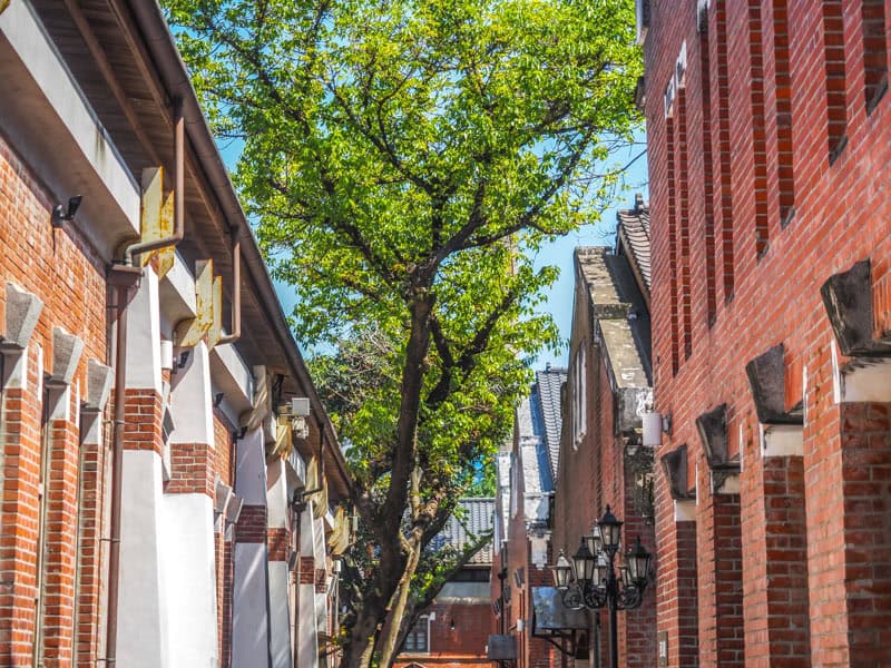 Looking down a lane between two rows of red brick buildings with some tall leafy trees growing in the middle