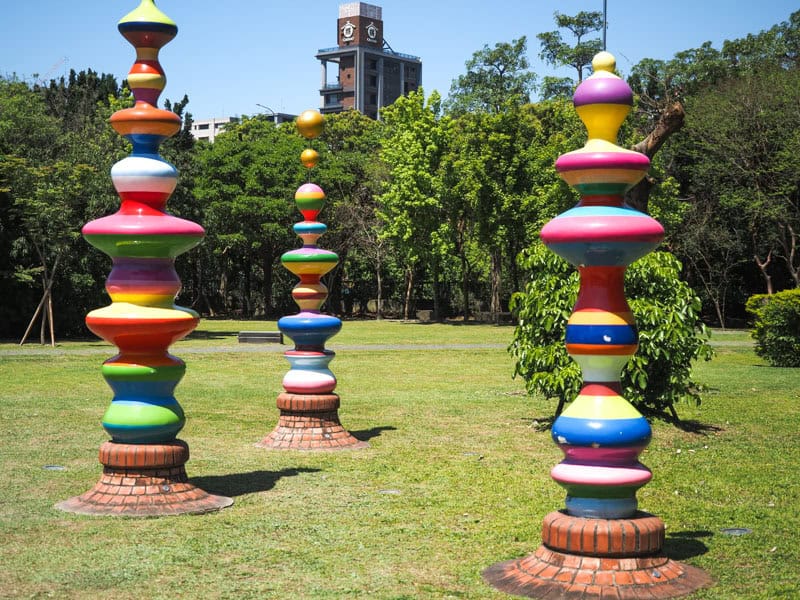 Three multi-colored tower sculptures in a grassy field with trees behind