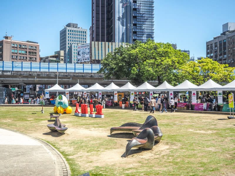 A grassy field with some artistic metal chairs and a row of white tents for some kind of market event, with buildings and elevated road in the background