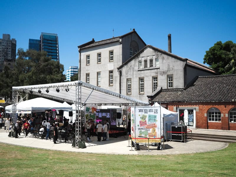 A grassy field, cement pad with an event tent and people under it, and some historic buildings of Huashan 1914 Creative Park behind it