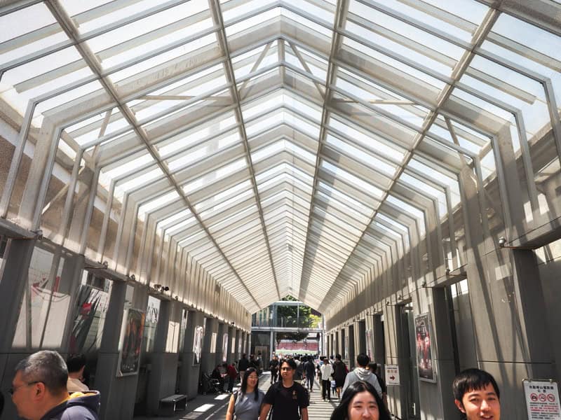 Looking down a long covered pedestrian walkway, with some people walking towards the camera, and an arched glass roof with metal beams