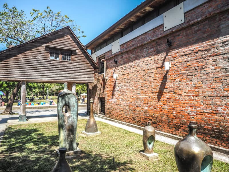 The side of a red brick building, with a lawn containing some sculptures beside it, and a unique pointed wooden building with no base connected to it at the back