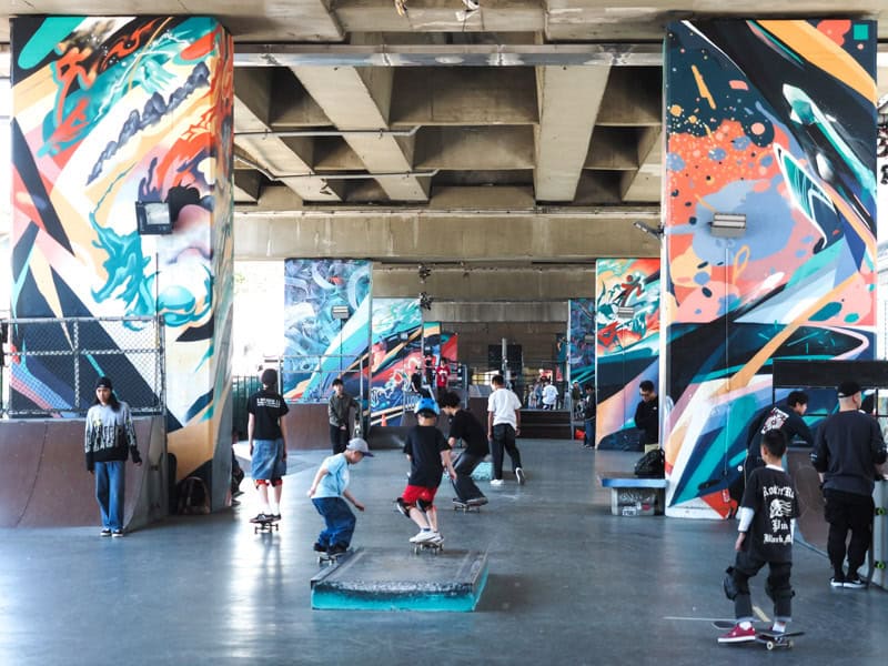 A skatepark under an elevated road, with street art on the columns and some kids skateboarding in the middle