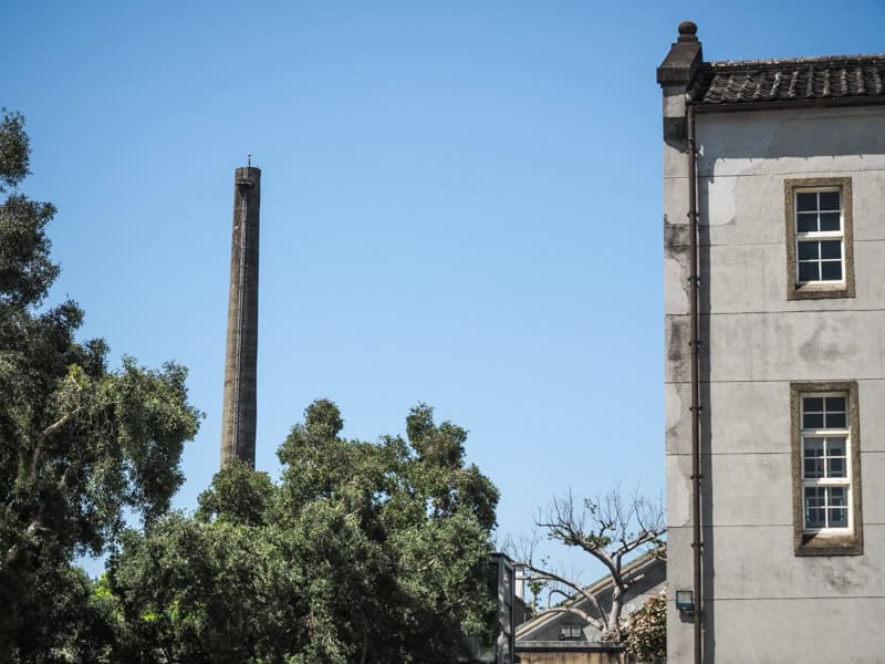 A cement smokestack rising above some trees on the left and the side of a tall cement building on the right